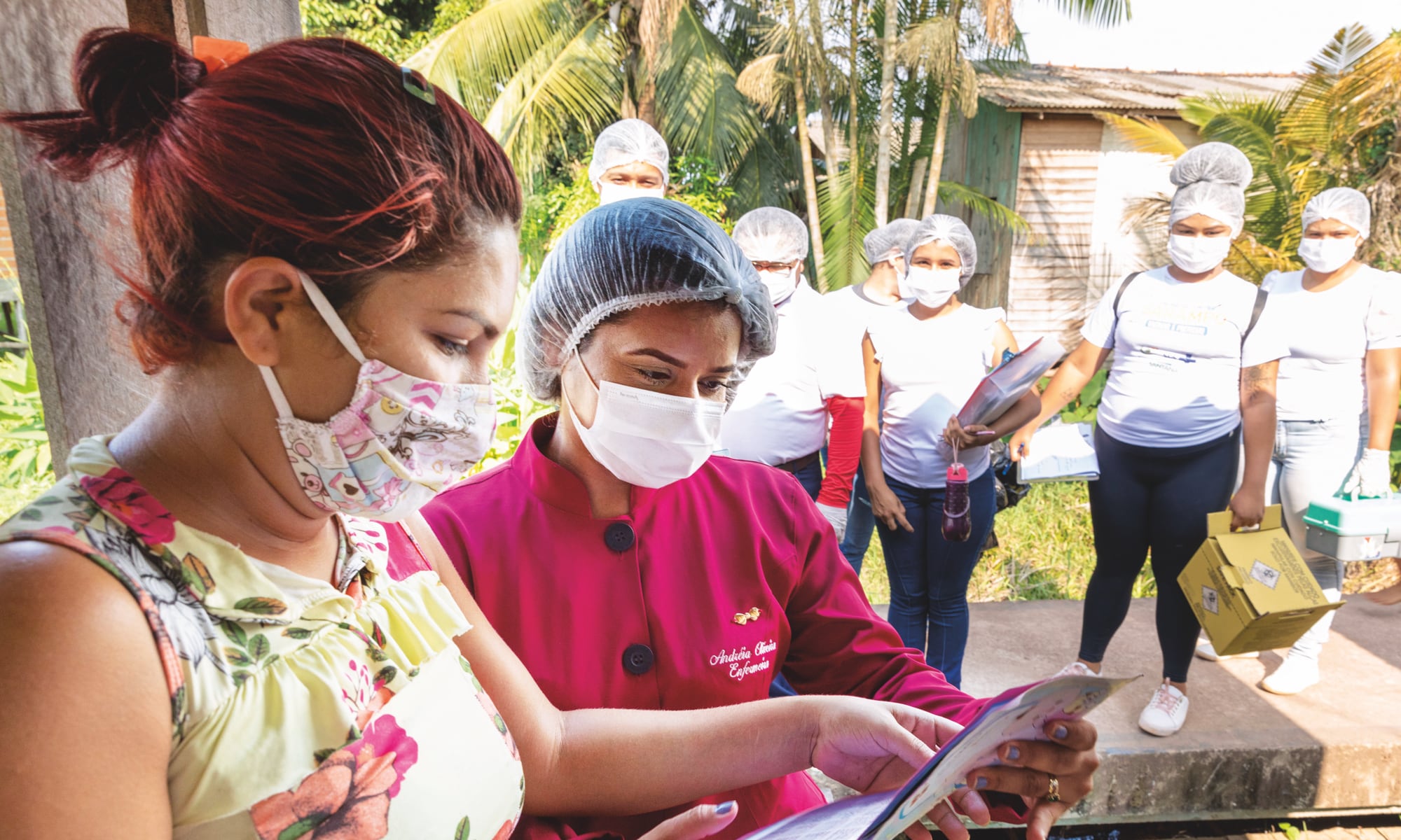 Two women wearing masks look at a document together, while several people in the background, also masked, stand and watch.