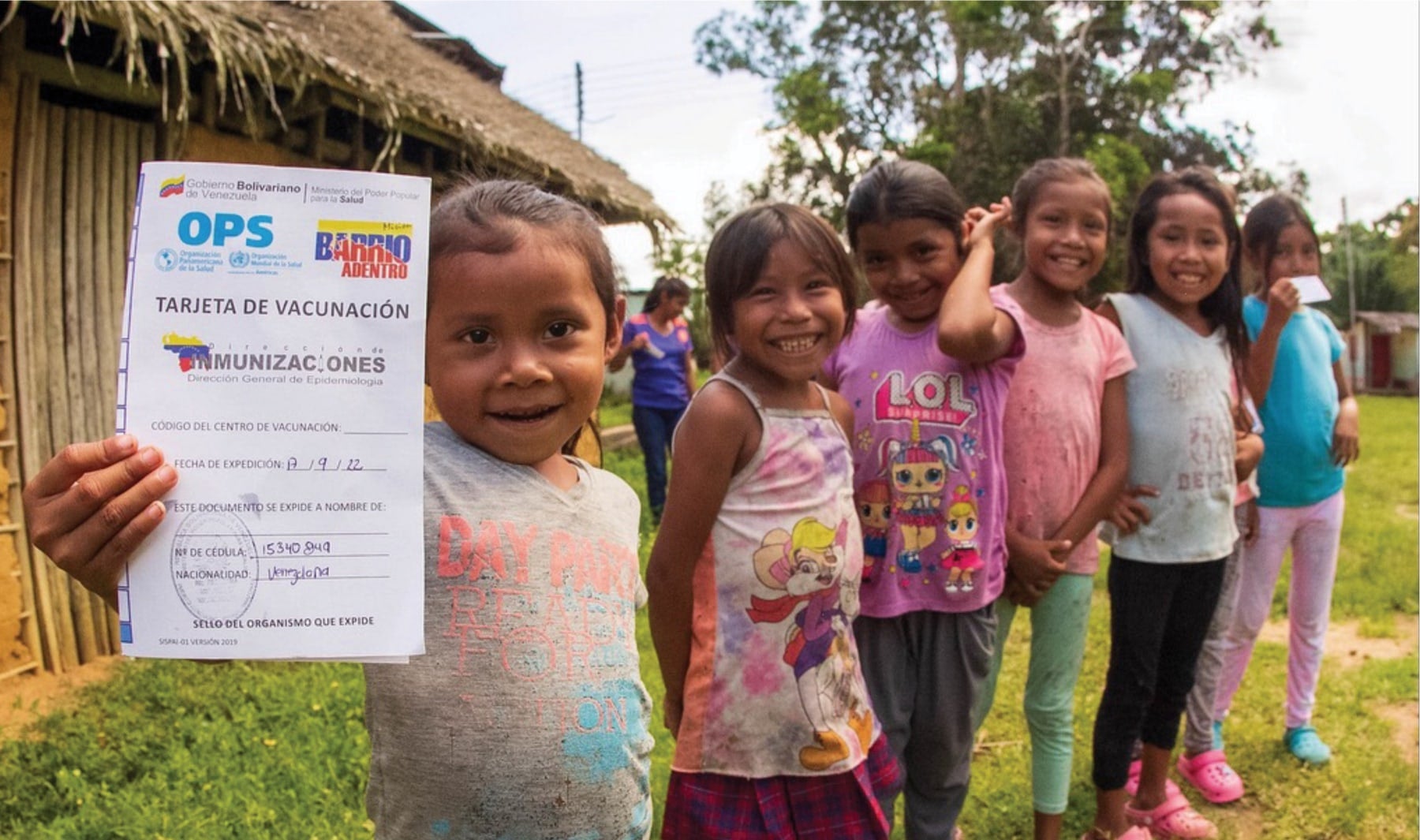 Smiling young girl holding a vaccination card while standing with a group of children outdoors.