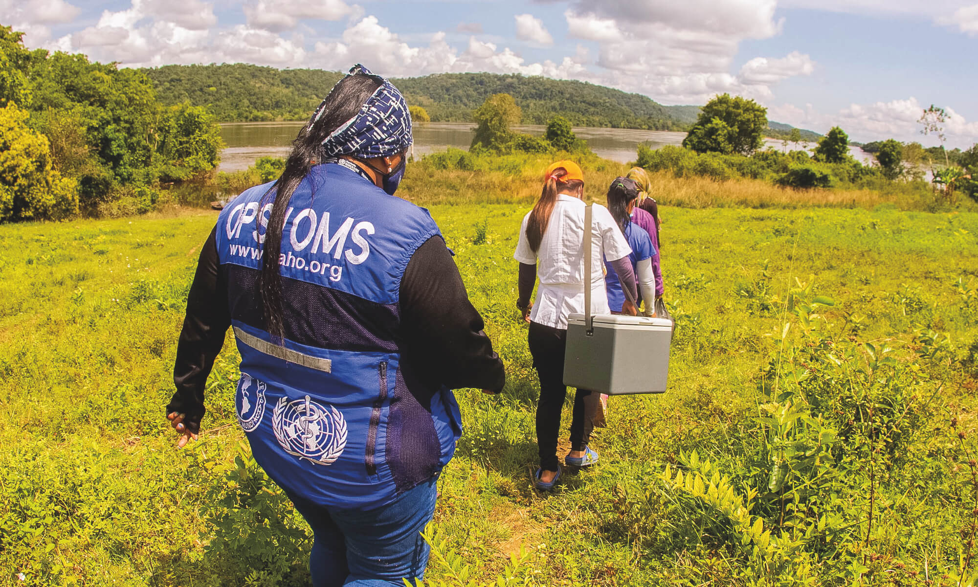 A group of health workers, one wearing a vest with PAHO and WHO logos, walk through a grassy field toward a river, carrying a medical cooler.