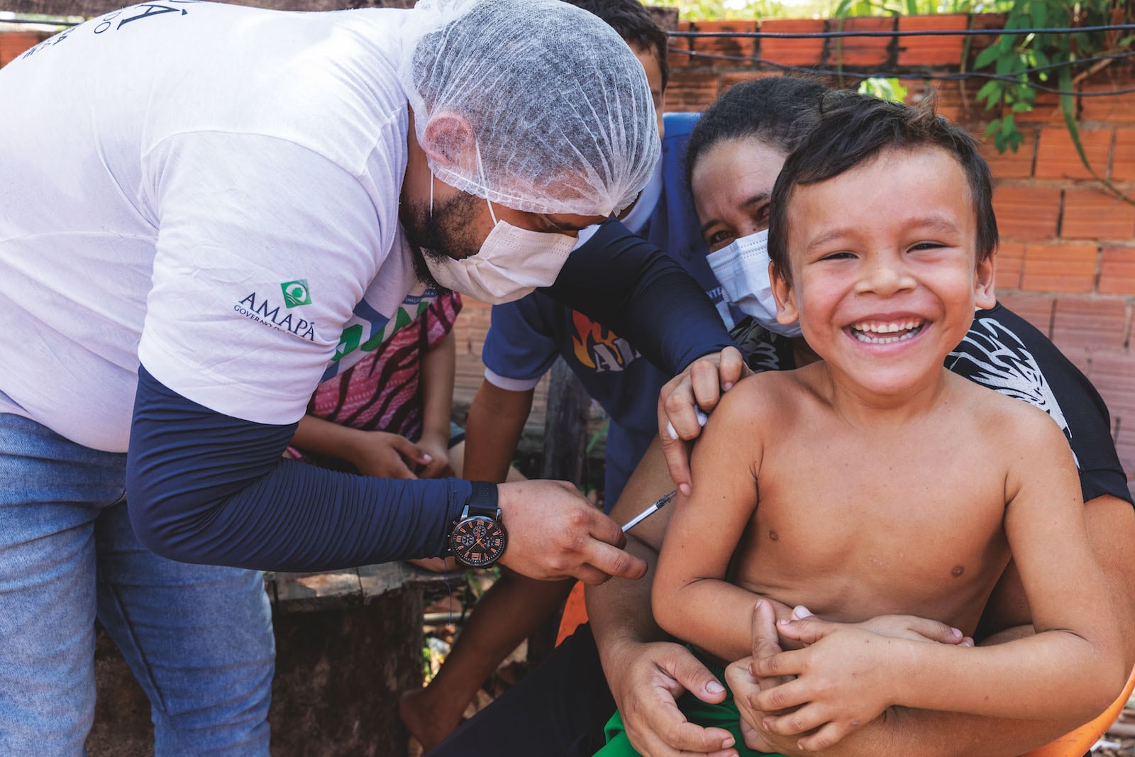 A healthcare worker vaccinates a child outdoors while another person assists.