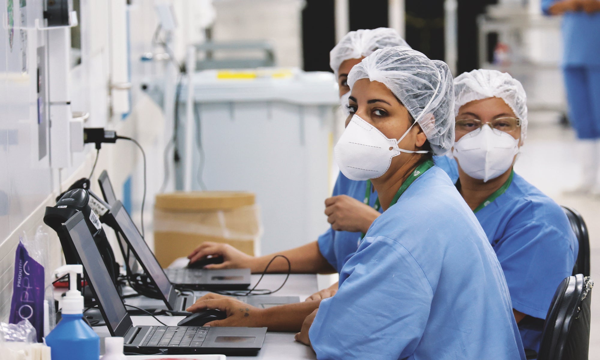 Healthcare workers in blue scrubs and masks work on laptops in a medical facility.