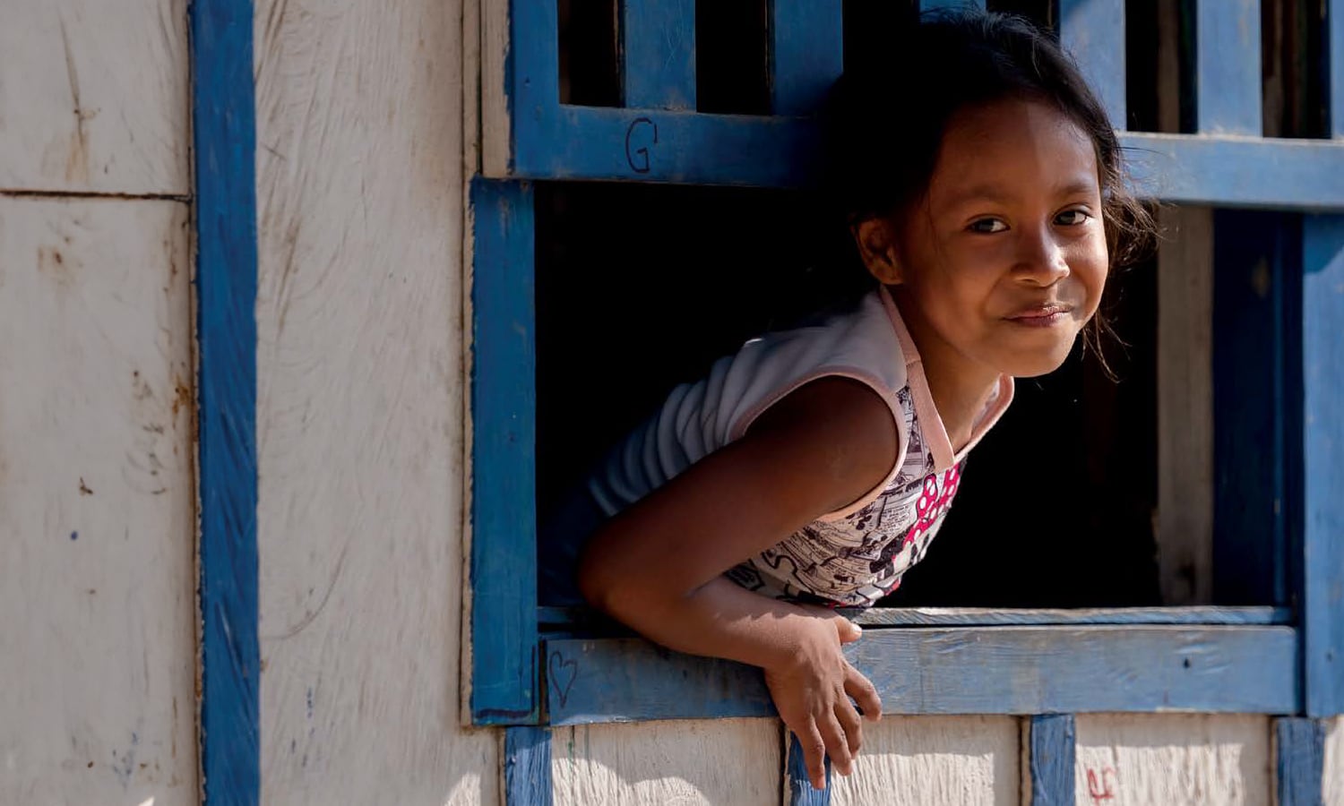 Young girl smiling while leaning out of a wooden window with blue trim.