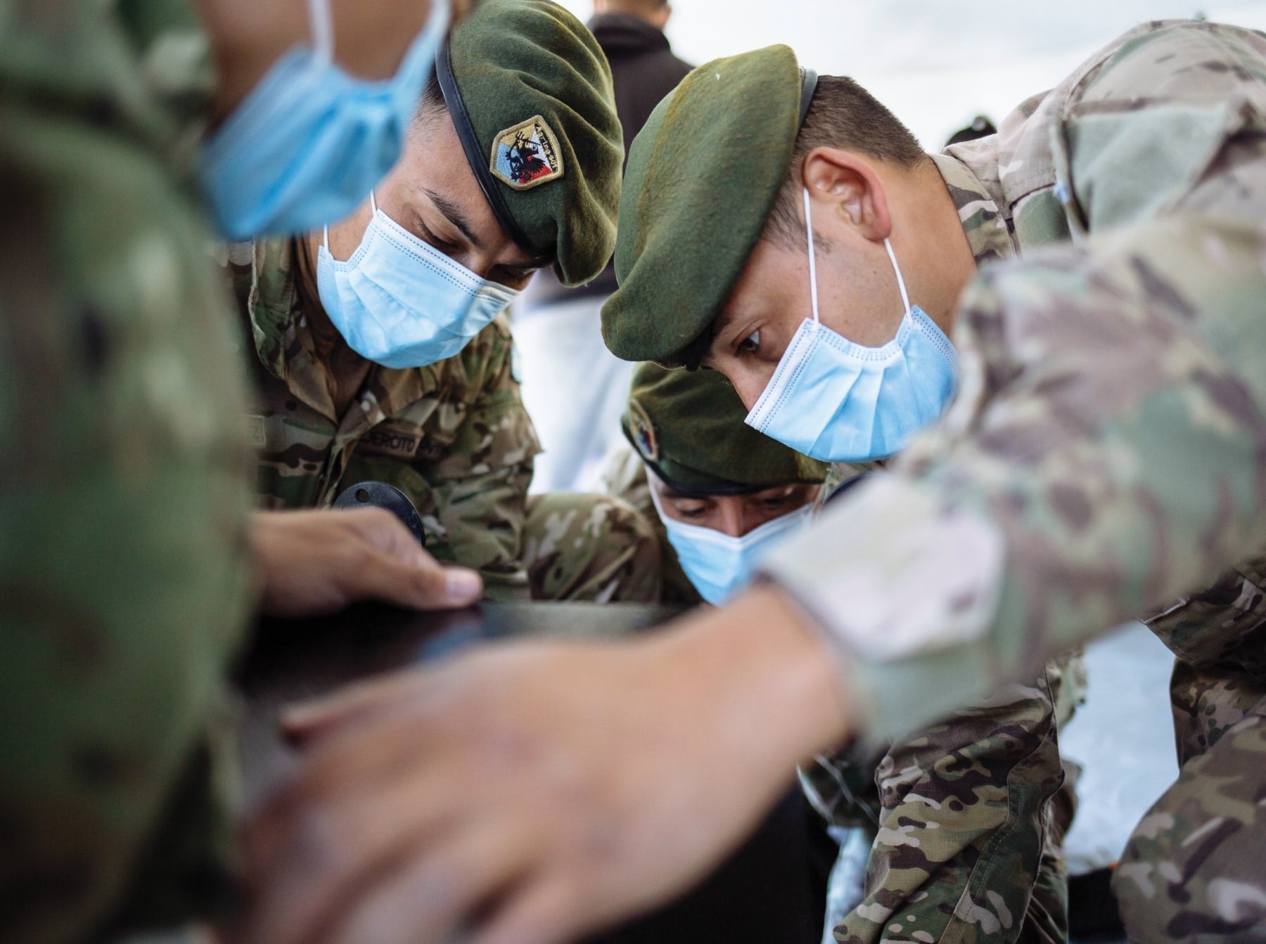 Soldiers in camouflage uniforms and face masks closely examine an object, appearing focused and collaborative.