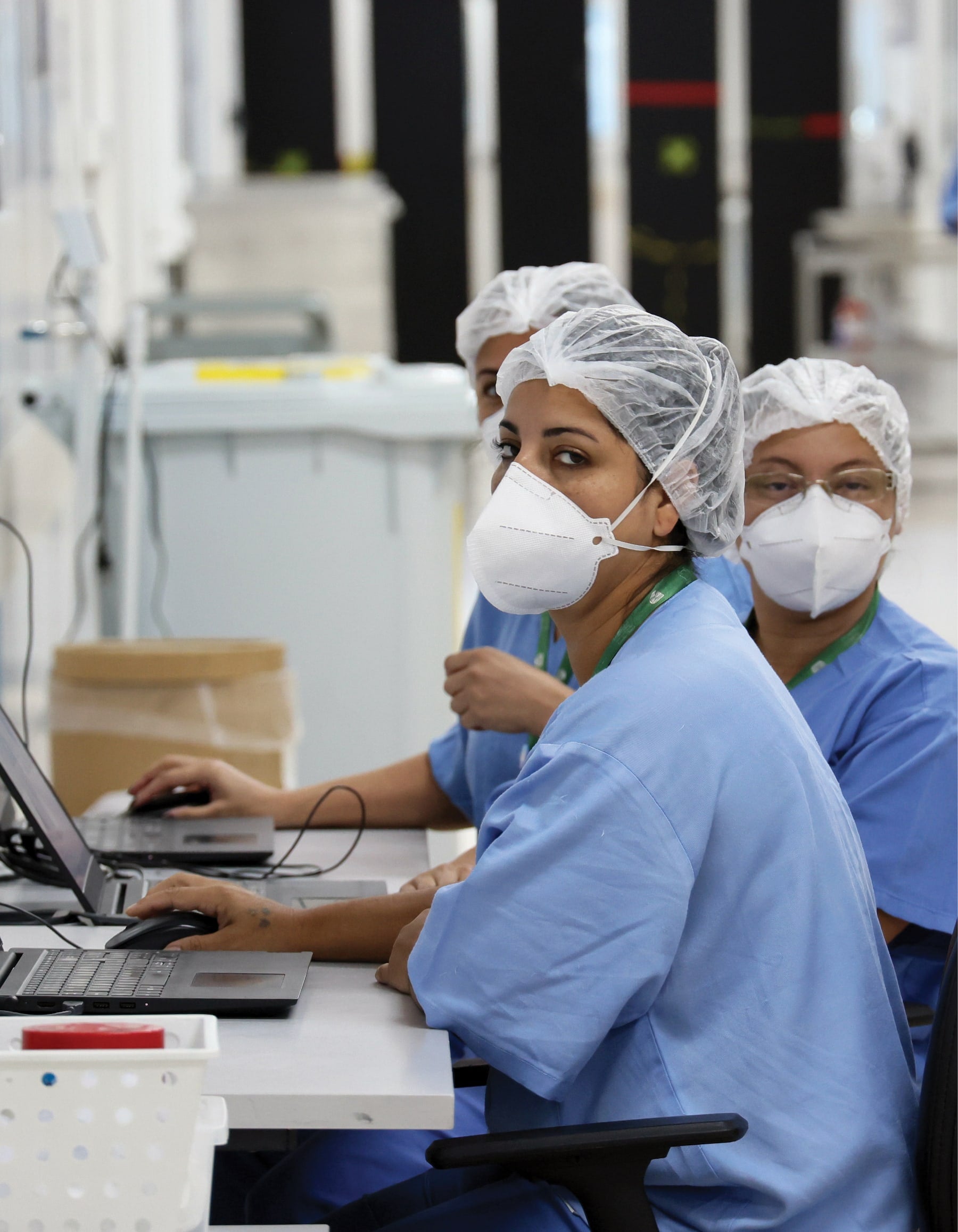 Healthcare workers in blue scrubs and masks work on laptops in a medical facility.