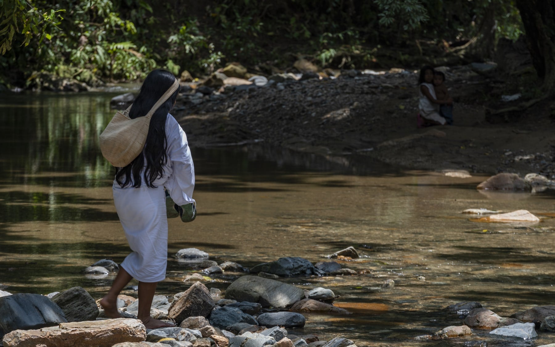 A person in white crossing a rocky stream with lush trees in the background.