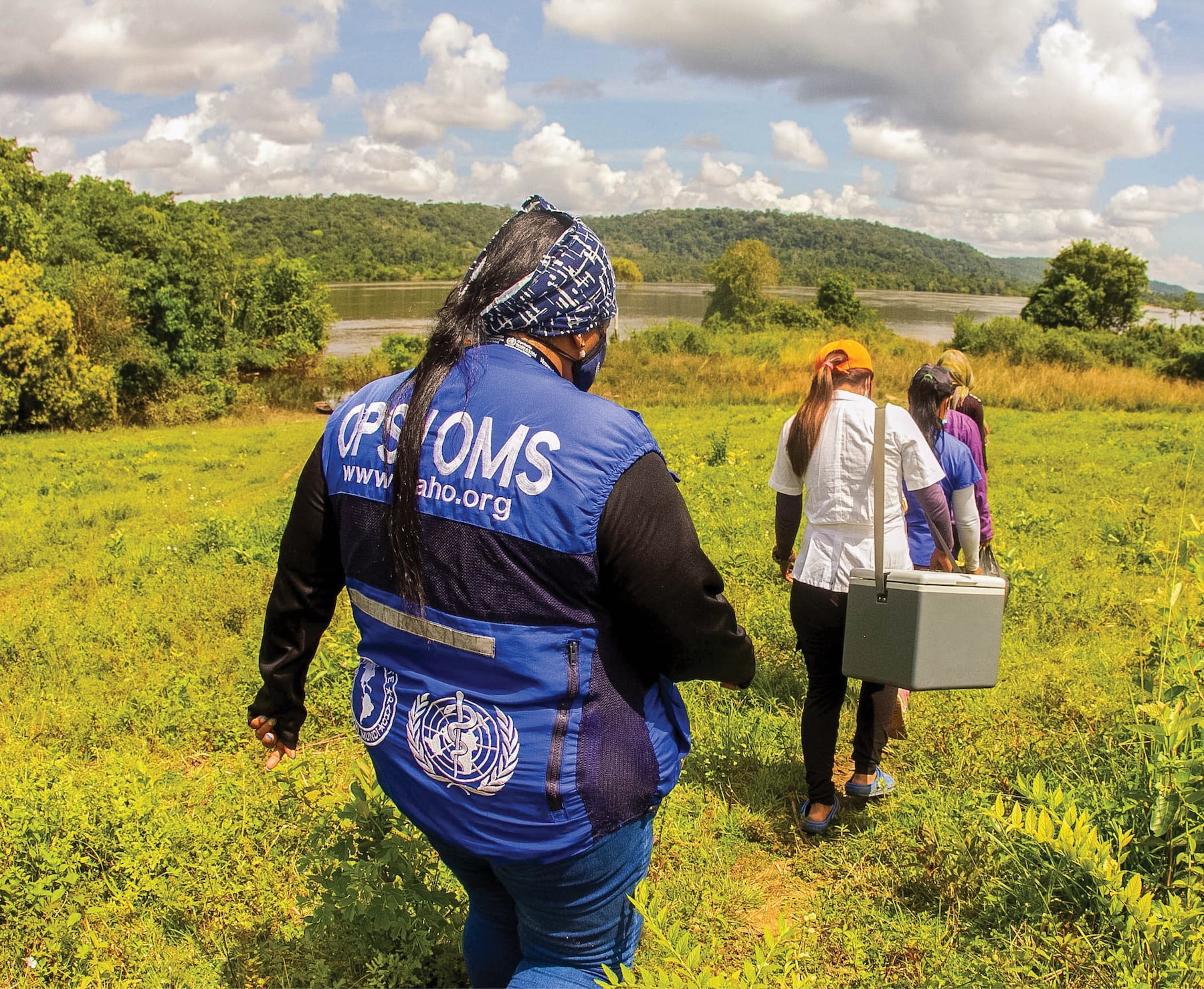 A group of health workers, one wearing a vest with PAHO and WHO logos, walk through a grassy field toward a river, carrying a medical cooler.