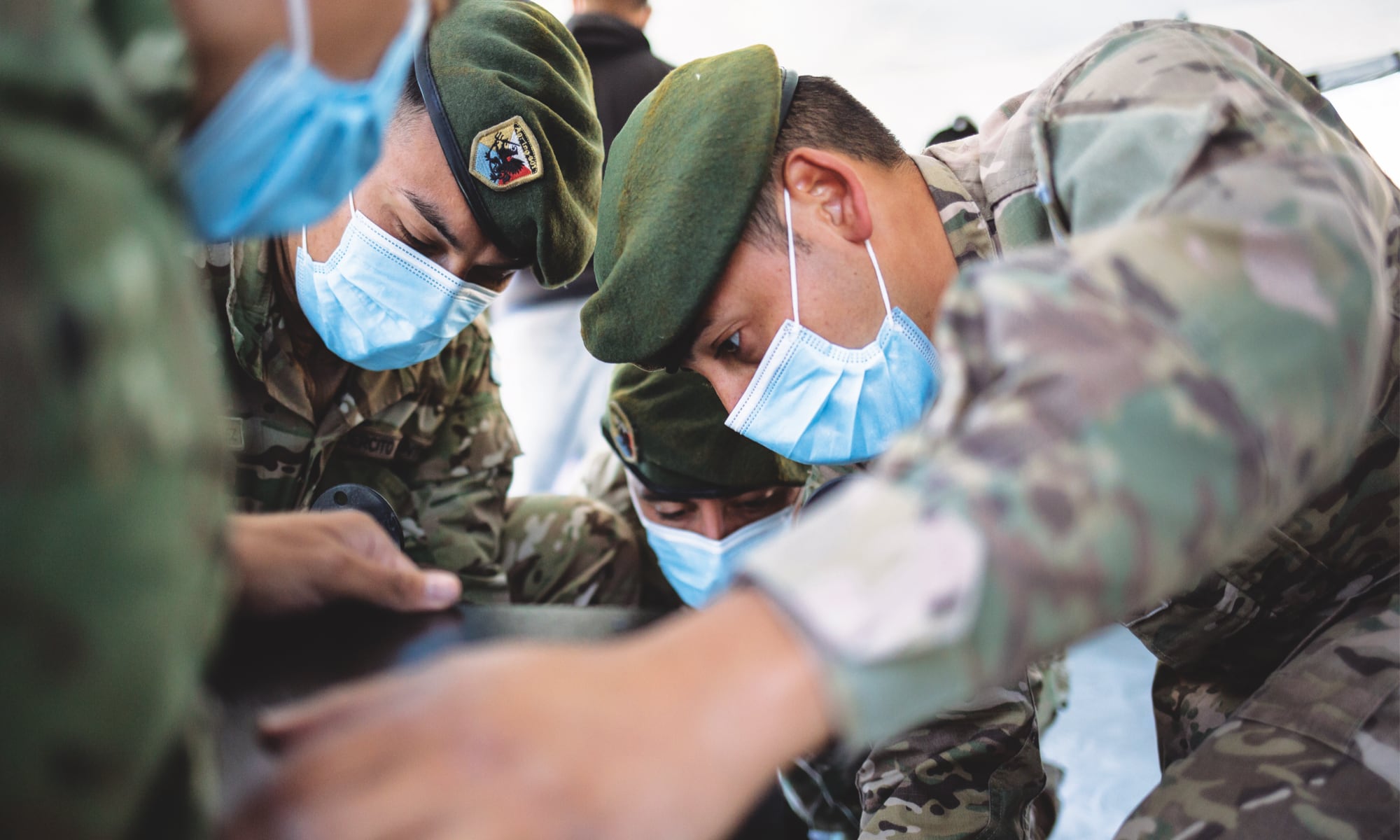 Soldiers in camouflage uniforms and face masks closely examine an object, appearing focused and collaborative.