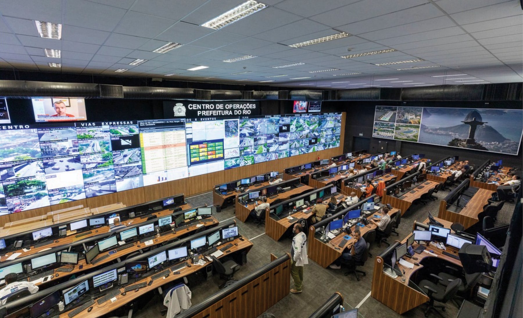 Modern control room with operators at workstations and large screens displaying data and video feeds.