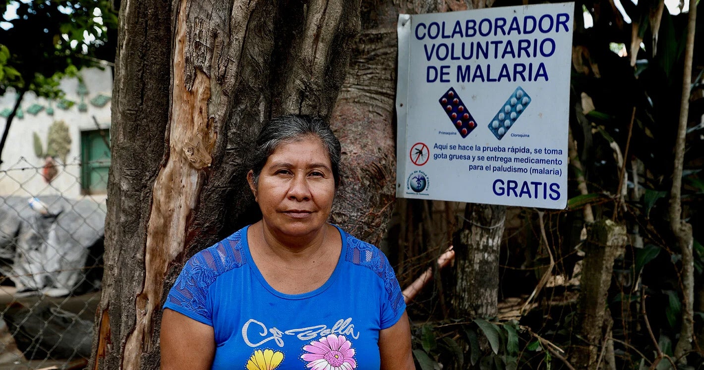 Imagen de una mujer de pie junto a un cartel que dice Colaborador Voluntario de Malaria