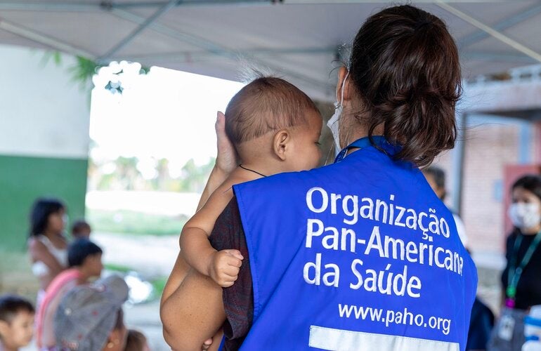 Mulher usando colete com o nome da OPAS, segura bebê yanomami