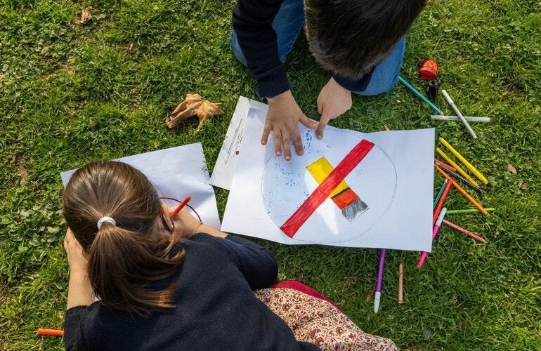 Two young children drawing anti tobacco signs over a grassy floor, surrounded by pens and pencils of may colors