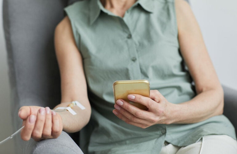 Mulher sentada em uma cadeira, recebendo medicação intravenosa e segurando o celular na outra mão.