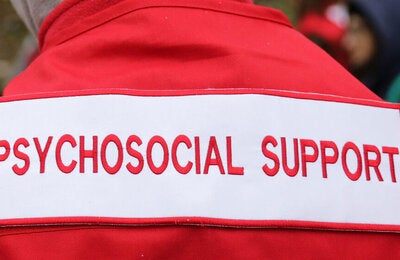 Photo of the back of a healthcare worker dressed with a red jacket with the sign "Psychosocial support" in red typography over white background. Only part of the back is seen, from the top of the shoulders to the mid part of the back