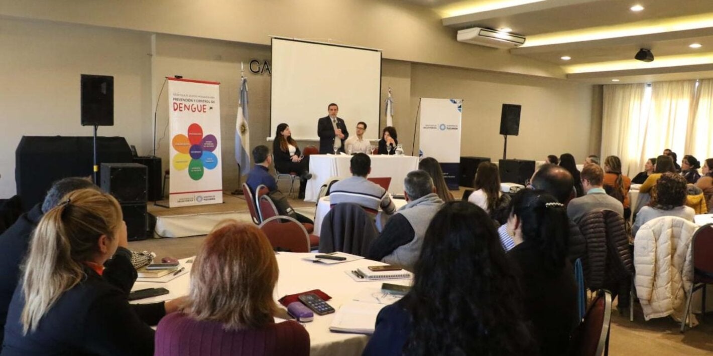 Momento de apertura de la capacitación. en primer plano se observan a los participantes de espaldas y hacia el fondo, el ministro de salud provincial y referentes de OPS y ministerio de salud de nación en la mesa de apertura. Un banner destaca: Estrategia de gestión integrada para la prevención y control del dengue. 