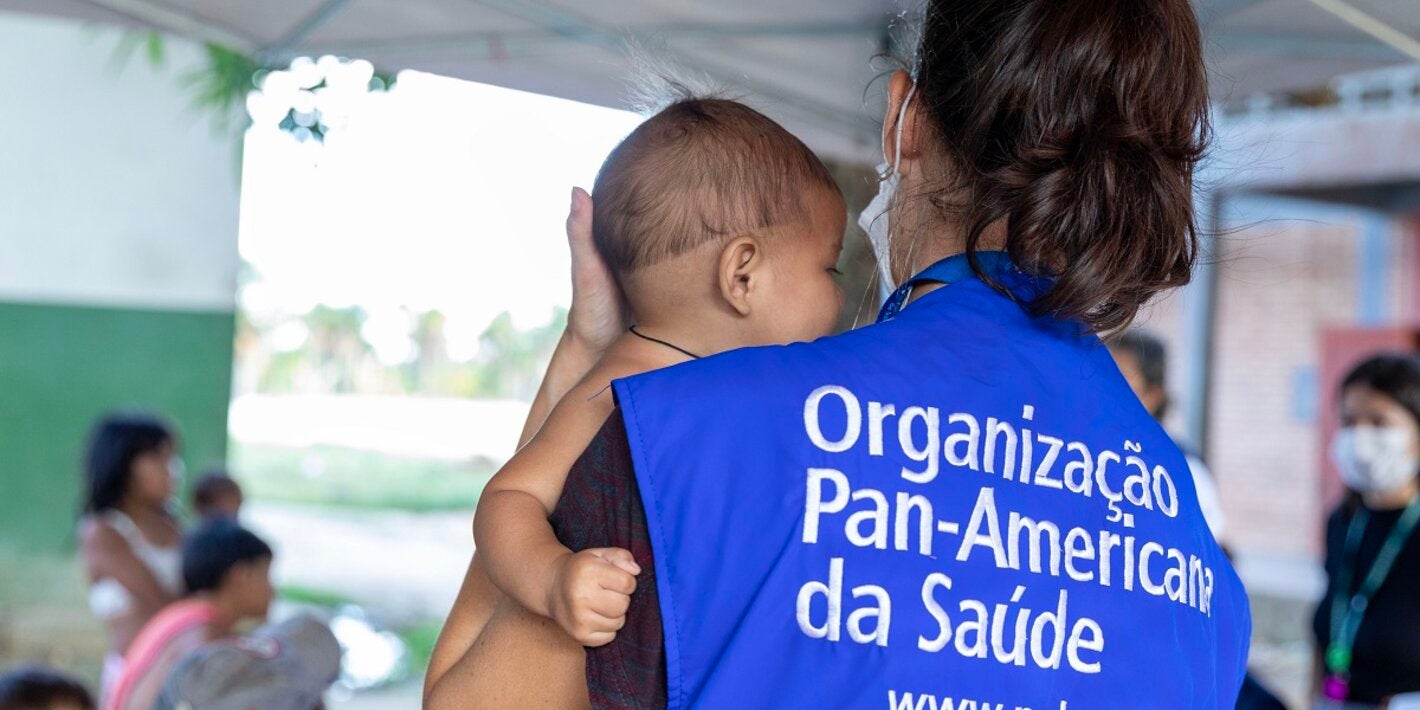 Mulher usando colete com o nome da OPAS, segura bebê yanomami