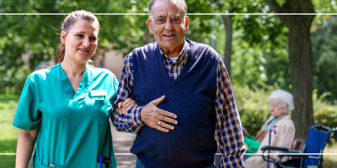 older man and nurse walking in wooded area with an older women in a wheelchair in the background