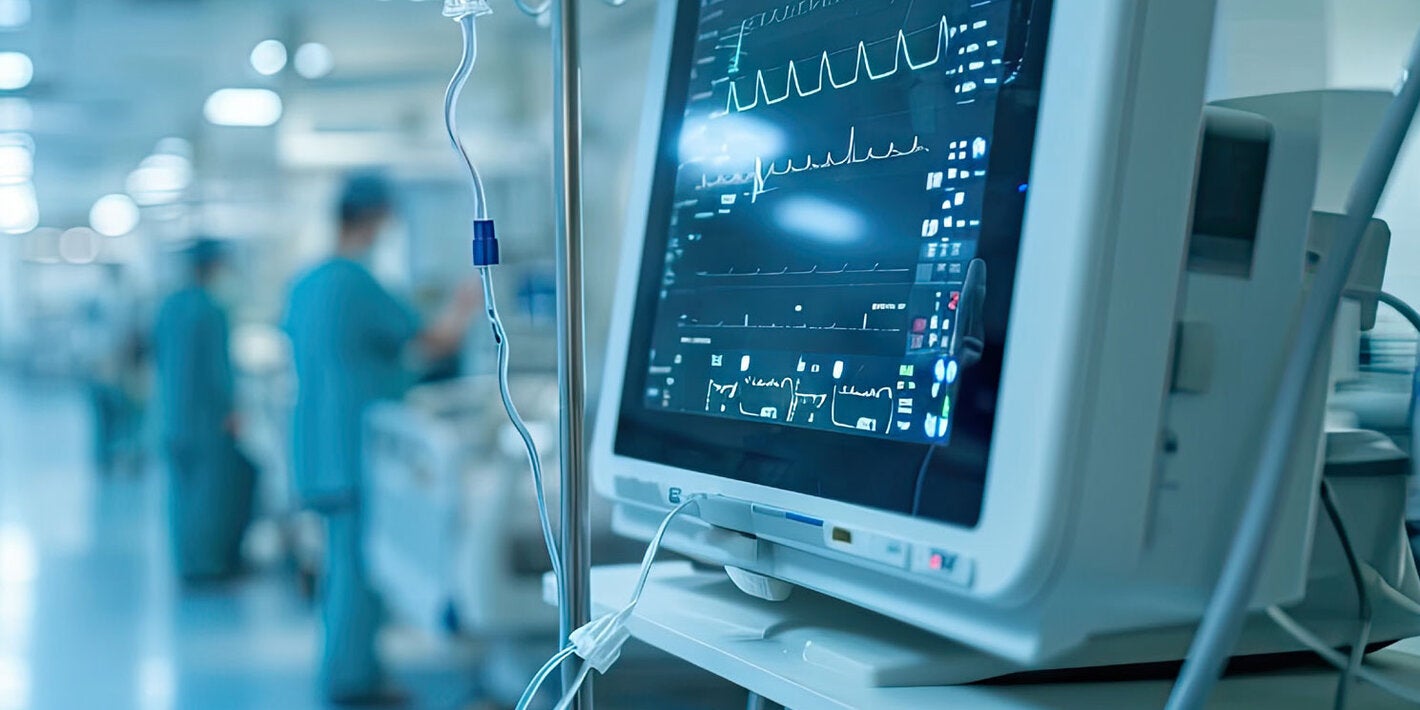 Hospital setting. The monitor with vital signs is in front. Behind are hospital beds and hospital staff.