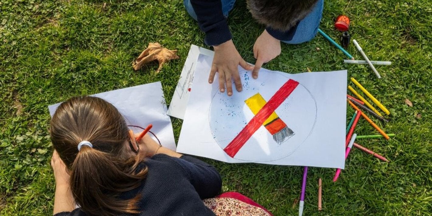 Two young children drawing anti tobacco signs over a grassy floor, surrounded by pens and pencils of may colors