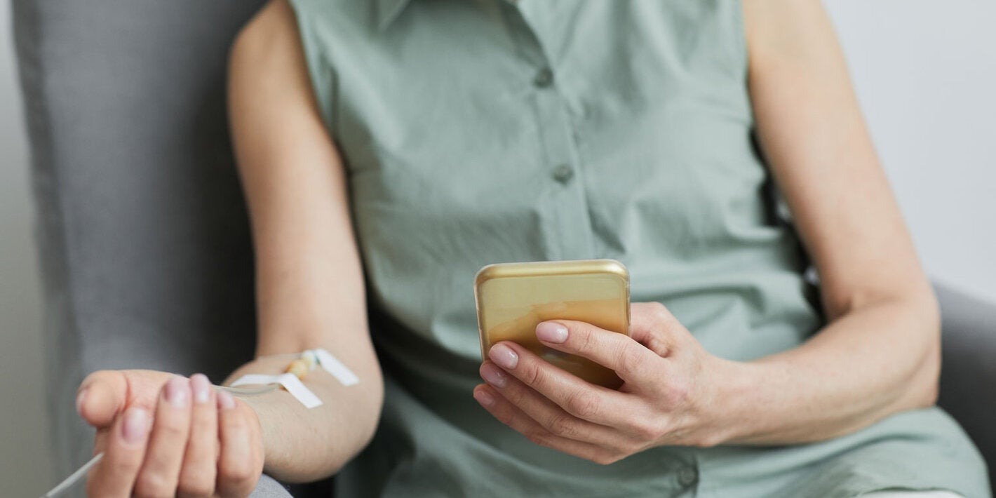 Mujer sentada en una silla, recibiendo medicación intravenosa y sosteniendo su teléfono celular en la otra mano.