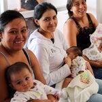 mujeres sentadas, mirando a cámara y sonriendo, con sus hijos bebés en brazos