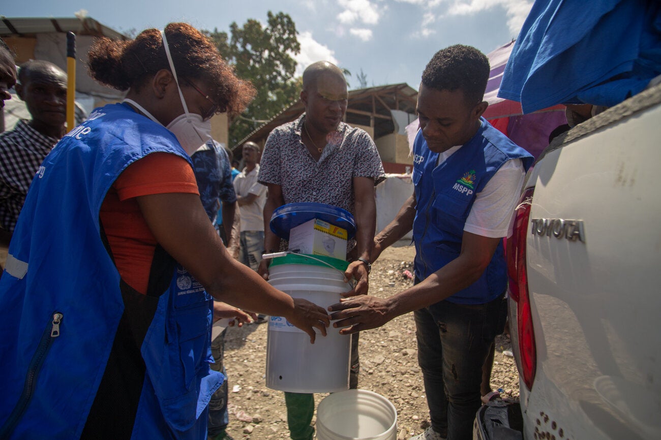 PAHO worker (woman) along with a Ministry of Health worker, both wearing blue vests, delivering supplies to a haitian man 