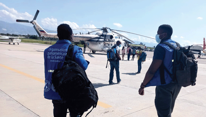 2 trabajadores de la OPS con chalecos azules y mochilas caminando hacia un helicóptero