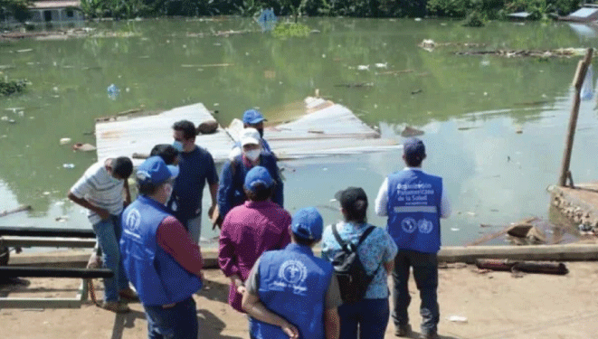 OPS y trabajadores comunitarios en la orilla de un río. Casas inundadas en la orilla opuesta 