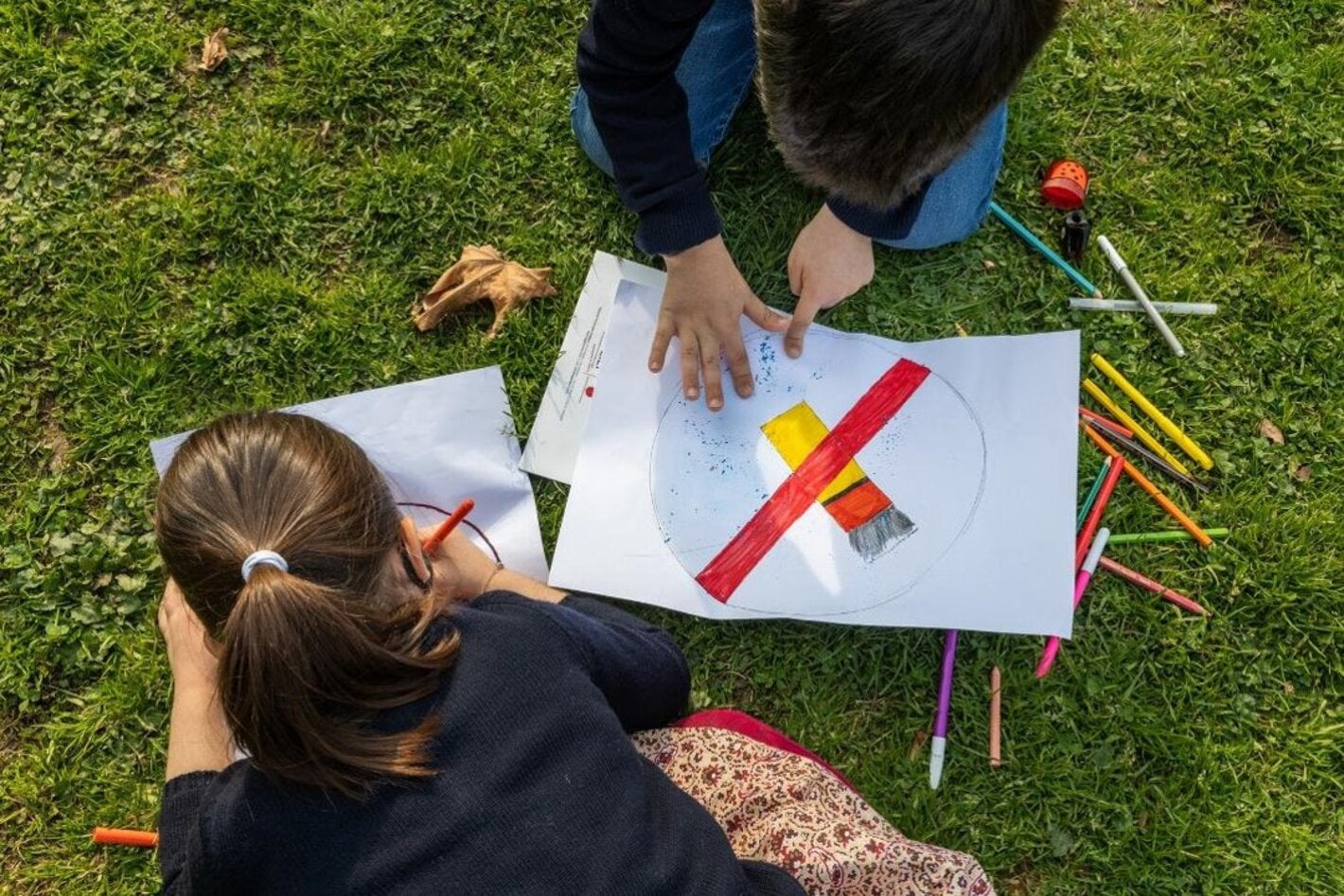 Two young children drawing anti tobacco signs over a grassy floor, surrounded by pens and pencils of may colors