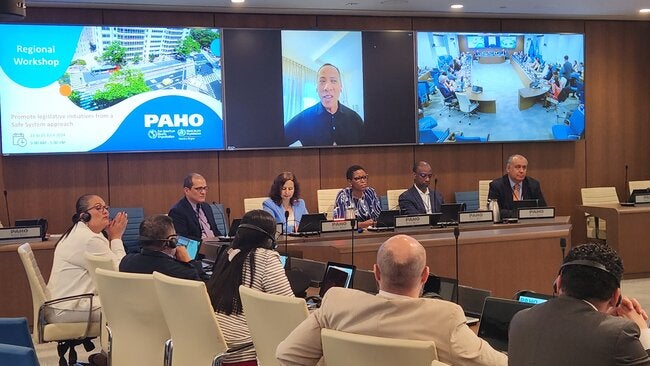 Photo of a meeting room with several people on a L shape display, with a speaker on a monitor in the center